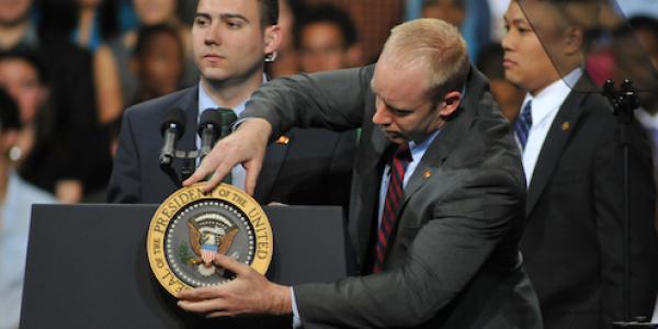 A man adjusts a symbol that says "President of the United States" on a podium while two other men in secret service garb look on.