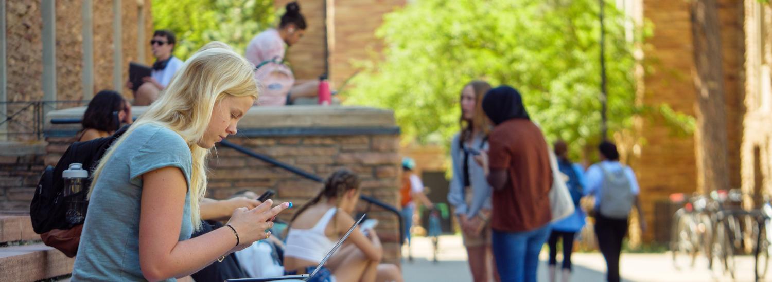 A student on campus looking at her phone with her laptop on her lap.